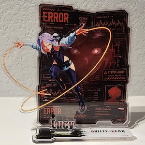 May include: An acrylic standee featuring a character with purple hair in a dynamic pose, holding a glowing orange wire. The background panel displays red digital interface graphics with the text ERROR, SYSTEM ALERT, and the name Lucy at the base.