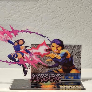 Könnte beinhalten: Ein klarer Acrylständer mit einer Comic-Illustration von Psylocke aus Marvel Super Heroes. Das Kunstwerk zeigt Psylocke in einem blau-violetten Kostüm mit rosa Energieeffekten. Auf der Basis des Displays steht "Psylocke" und "Marvel Super Heroes."