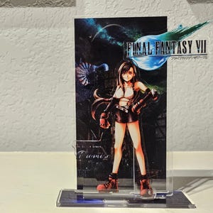 May include: A clear acrylic standee featuring Tifa Lockhart from Final Fantasy VII. The character wears a white tank top, black skirt, and red boots. The background panel displays the Final Fantasy VII logo and a dark, atmospheric cityscape scene.