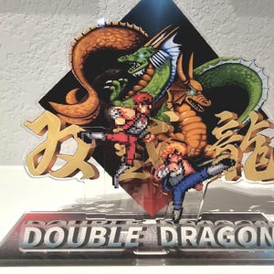 May include: A clear acrylic display featuring pixelated characters and dragons from the video game Double Dragon. The design includes the game's title and Japanese characters. The dragons are in shades of green, orange, and brown.