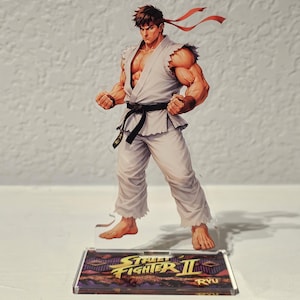 Può includere: Una statuetta in acrilico di Street Fighter II di Ryu in un gi bianco con cintura nera e fascia rossa. La statuetta è su una base rettangolare con il logo del gioco e il nome del personaggio.