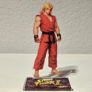 May include: A standing figure of Ken from Street Fighter II, in a red martial arts uniform with a black belt and wristbands. The character has blonde hair and is posed on a clear base with the game's logo and the name "Ken".