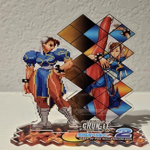 May include: A clear acrylic figure of Chun-Li from SNK vs. Capcom 2. The figure depicts two versions of Chun-Li in her blue dress. The base features the game's logo and text. The figure is a collectible item.