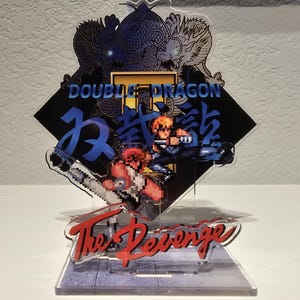 May include: A clear acrylic display featuring pixelated characters from the video game "Double Dragon." The design includes the game's title, Japanese characters, and the phrase "The Revenge" in red. The display is on a clear base.