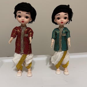 May include: Two dolls dressed in traditional Indian attire. One doll wears a maroon and gold outfit, the other a green and gold outfit. Both dolls have black hair, red lips, and white shoes. The dolls' outfits include a top and a dhoti-style bottom.