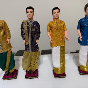 May include: Four male fashion dolls dressed in traditional Indian attire. The dolls wear various outfits, including gold, blue, and green garments with intricate embroidery and embellishments. Each doll stands on a small, decorative platform.