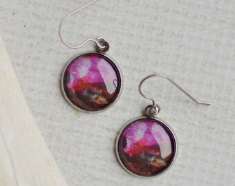 May include: A pair of round, silver-toned earrings with a colorful, abstract design. The earrings feature a vibrant mix of pink, purple, and brown hues. The earrings are displayed on a light-colored surface next to a piece of driftwood.