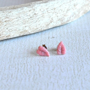 May include: A pair of small, pink leaf-shaped stud earrings. The earrings are set against a light background with a piece of driftwood in the upper portion of the image. The earrings are a delicate accessory.