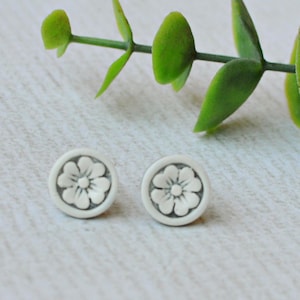 May include: A pair of small, circular stud earrings featuring a raised, white floral design centered on a light gray background. The earrings have a matte finish and a simple, clean aesthetic, showcasing a detailed flower petal pattern within each circular frame.
