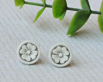 May include: A pair of small, circular stud earrings featuring a raised, white floral design centered on a light gray background. The earrings have a matte finish and a simple, clean aesthetic, showcasing a detailed flower petal pattern within each circular frame.