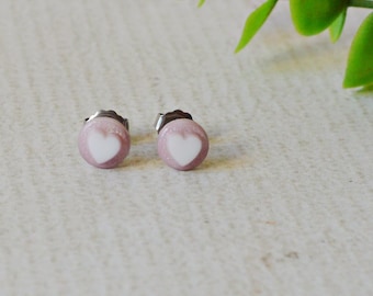 May include: A pair of small, round stud earrings. Each earring is light pink with a white heart design. A green leafy sprig is in the background. The earrings are likely made of metal and are a jewelry item.