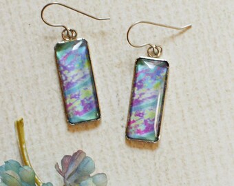 May include: A pair of rectangular dangle earrings with a colorful, abstract design in shades of blue, purple, and green. The earrings have silver-colored hooks. A small, decorative floral element is also present.