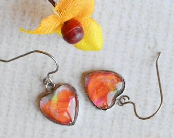 May include: A pair of heart-shaped drop earrings featuring a vibrant orange, yellow, and red marbled pattern encased in a thin metal frame with silver-toned hook wires, positioned near a small yellow flower with a dark brown center.