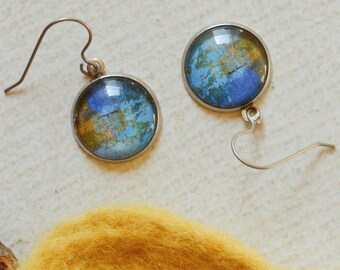 May include: A pair of round dangle earrings with a blue and gold design, set against a light background. A yellow, leaf-shaped felted wool object and a small twig are also present.