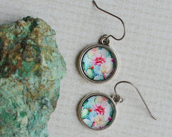 May include: A pair of round silver-toned drop earrings featuring a colorful floral pattern with pink, blue, and yellow blossoms set under a clear dome, accompanied by a rough, textured piece of green mineral stone with brown inclusions.