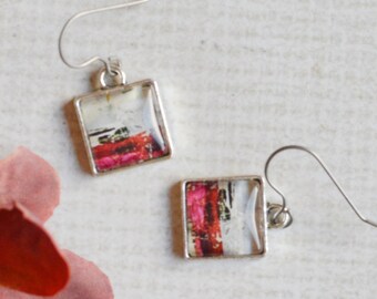 May include: A pair of silver-toned square drop earrings featuring a graphic print with red, white, and black abstract patterns, suspended from curved silver-toned ear wires, accompanied by a single artificial flower with red and pink petals.