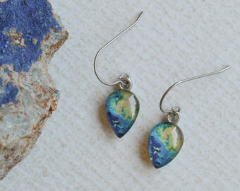May include: A pair of teardrop-shaped earrings with a blue, green, and yellow marbled design. The earrings hang from silver-colored hooks. A rough, textured rock with blue and brown patches sits to the left of the earrings on a white surface.