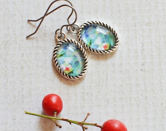May include: A pair of oval-shaped drop earrings featuring a silver-toned twisted metal border and a colorful floral pattern under a glass cabochon, accompanied by a small twig with three bright red berries positioned below the jewelry.