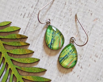 May include: A pair of teardrop-shaped earrings with green and blue accents, next to a green and yellow leaf-shaped wooden cutout. The earrings have silver-colored hooks. The leaf cutout is a detailed representation of a fern frond.