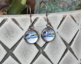 May include: A pair of round drop earrings featuring a silver-toned metal setting and a small circular insert with an abstract blue, white, and yellow painted design. The earrings hang from thin, curved silver-toned metal ear wires.