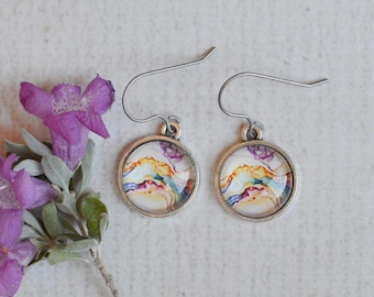 May include: A pair of round silver-toned drop earrings featuring a colorful abstract pattern with swirls of blue, orange, and purple. The earrings have curved metal hooks and are displayed next to a small sprig of purple flowers with green leaves.