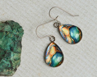 May include: A pair of teardrop-shaped earrings featuring a swirling abstract pattern of blue, orange, and gold colors, suspended from silver-toned metal hooks, positioned next to a rough, textured piece of green mineral stone.