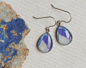 May include: A pair of teardrop-shaped earrings featuring a marbled pattern of blue, white, and purple tones set in dark metal frames with hook-style wires, positioned next to a rough, textured rock with blue and brown mineral deposits.