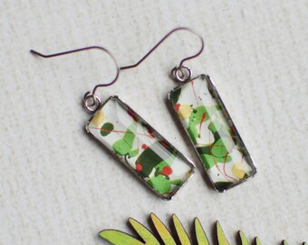 May include: A pair of rectangular earrings with a botanical design, featuring green and red accents, hang from silver hooks. A green and yellow leaf is positioned below the earrings on a white surface. The earrings are a unique accessory.