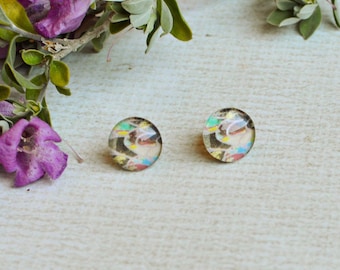 May include: A pair of round stud earrings featuring a multi-colored abstract pattern with shades of blue, yellow, and black under a clear glass dome. The earrings are positioned next to a sprig of small purple flowers with green leaves.