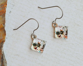 May include: A pair of dangle earrings featuring small, square-shaped charms with a clear, glossy finish. The charms contain a multi-colored, speckled pattern with hints of green, red, and black. Each charm hangs from a thin, dark metal hook.