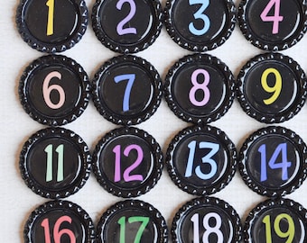 May include: A collection of thirty-one black bottle caps arranged in a grid, each featuring a single number from one to thirty-one printed in a variety of bright colors including yellow, purple, blue, pink, green, orange, and light red.