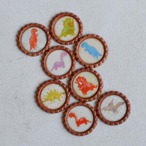 May include: Eight brown round bottle caps with different dinosaur designs. The dinosaurs are in various colors, including red, blue, green, and purple.
