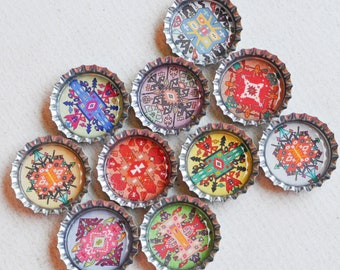 May include: Ten colorful bottle caps with silver rims and intricate patterned designs. The bottle caps are arranged in a circular pattern on a white surface.