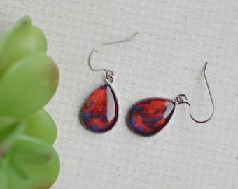 May include: A pair of teardrop earrings with a red and blue marbled design. The earrings have silver-colored hooks and are set against a white background. A green plant is in the background.