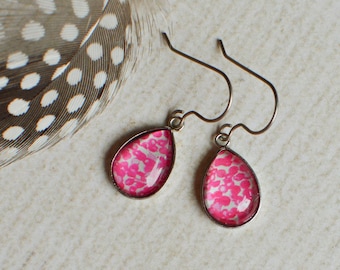 May include: A pair of teardrop-shaped earrings with a pink floral pattern set in a silver-colored metal. The earrings are displayed with a speckled feather.