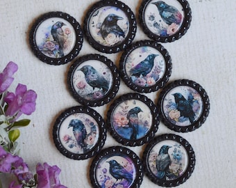 May include: Ten circular black bottle cap ornaments featuring printed illustrations of black birds perched among colorful floral arrangements, arranged in a cluster next to a sprig of purple flowers with green leaves and stems.