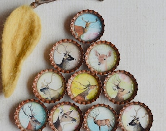 May include: A collection of ten bottle caps with deer illustrations, arranged in a triangle. Each cap features a different deer image with watercolor-style backgrounds. A yellow felt leaf and a small twig are also present.