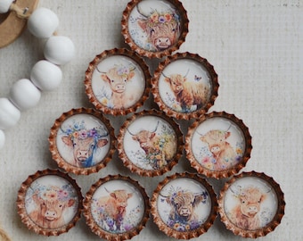 May include: A collection of bottle caps with watercolor illustrations of Highland cows. Each cap features a different cow with floral accents. The caps are arranged in a triangular shape on a white surface.