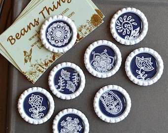 May include: A collection of navy blue and white decorative magnets with various floral and geometric designs. The magnets are made from bottle caps. A business card with the text "Beans Things" is also present.