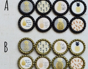 May include: A collection of bottle cap magnets with various designs. Some feature a pineapple, others have text like "Life is Sweet" and "You are the pina to my colada." The caps have black or gold rims.