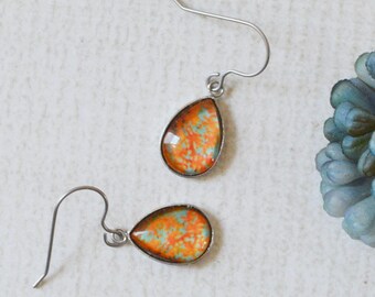 May include: A pair of teardrop-shaped dangle earrings featuring a vibrant orange and yellow marbled pattern set in a thin silver-toned metal frame with curved ear wires, positioned next to a small cluster of blue-grey flower petals.