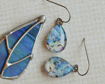 May include: A pendant and a pair of earrings. The pendant is a blue and silver teardrop shape. The earrings are teardrop-shaped with a blue and white pattern. The jewelry is set against a neutral background.
