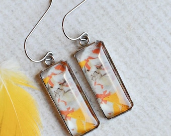 May include: A pair of rectangular drop earrings featuring a metallic frame and a multi-colored inlay of white, orange, and grey abstract patterns, accompanied by a single small yellow feather with a white tip positioned next to the jewelry.