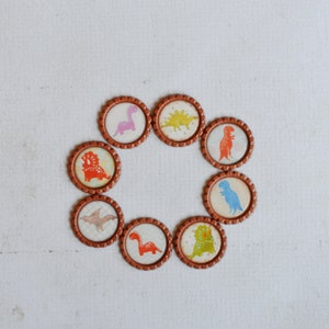 May include: Eight brown bottle caps with different dinosaur illustrations. The dinosaurs are in various colors, including pink, green, blue, and orange.