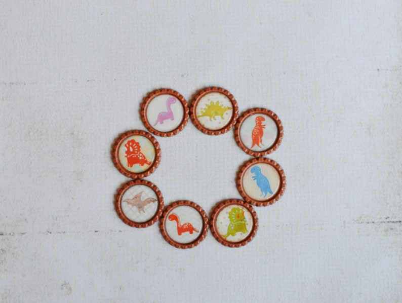 May include: Eight brown bottle caps with different dinosaur illustrations. The dinosaurs are in various colors, including pink, green, blue, and orange.