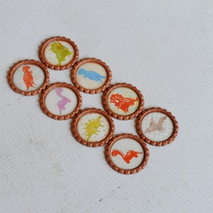 May include: Eight brown bottle caps with different dinosaur designs. The dinosaurs are in various colors, including orange, pink, blue, green, and yellow.