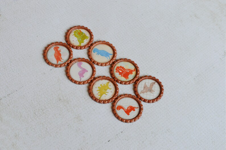 May include: Eight brown bottle caps with different dinosaur designs. The dinosaurs are in various colors, including orange, pink, blue, green, and yellow.