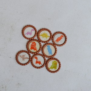 May include: Eight round brown bottle caps with different dinosaur designs. The dinosaurs are in various colors, including pink, orange, blue, green, and yellow.