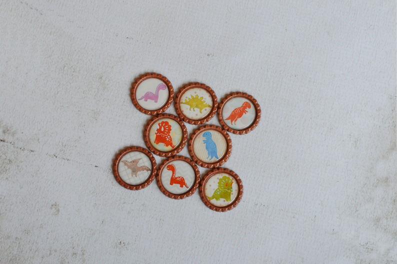 May include: Eight round brown bottle caps with different dinosaur designs. The dinosaurs are in various colors, including pink, orange, blue, green, and yellow.