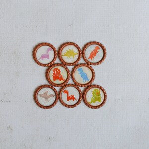 May include: Nine brown round bottle caps with different dinosaur designs. The dinosaurs are in various colors, including pink, orange, blue, green, and yellow.